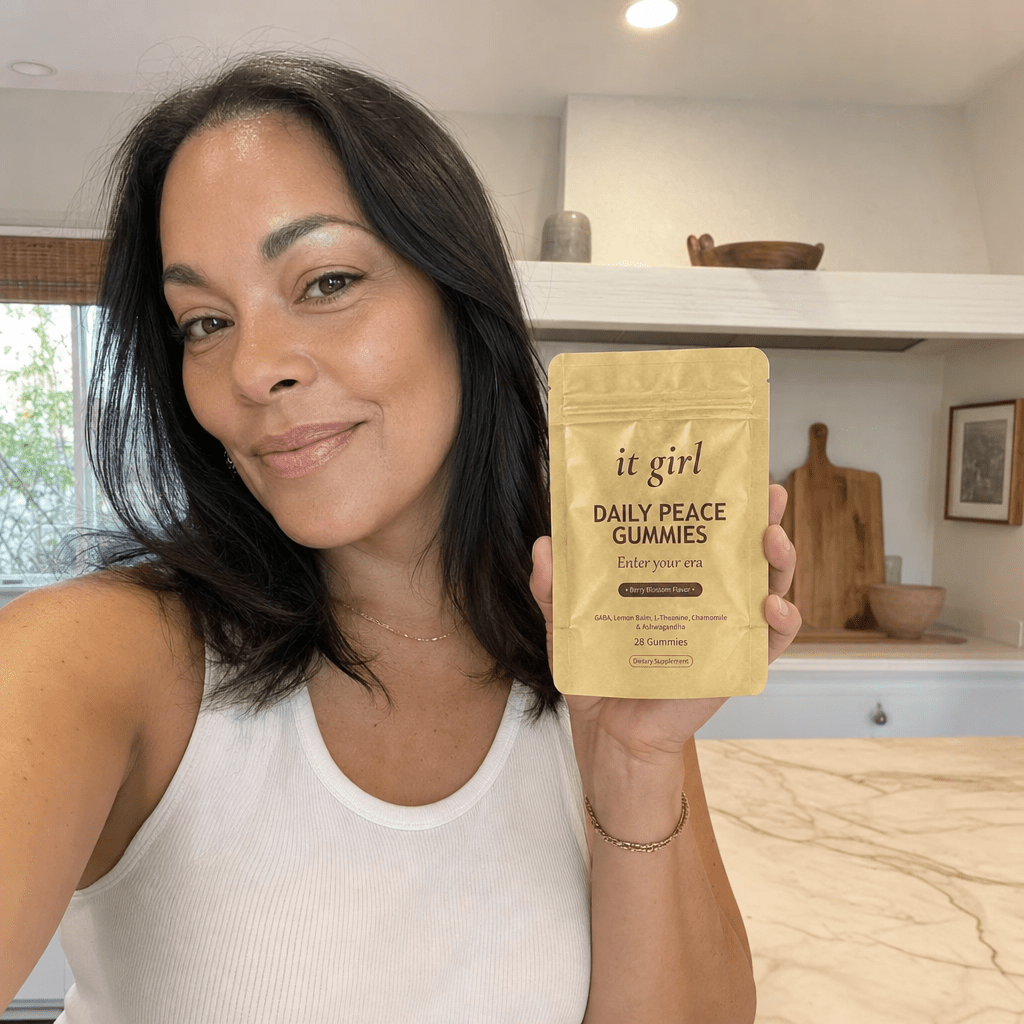 A capable woman in her forties in her warm early-evening kitchen holding the gold It Girl Daily Peace pouch with a relieved calm expression, no wine glass