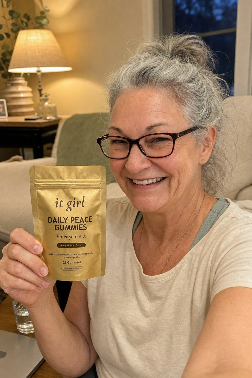 Warm candid selfie of a calm, composed woman in her early sixties in a cozy evening living room, holding up the gold It Girl Daily Peace pouch, no wine in frame