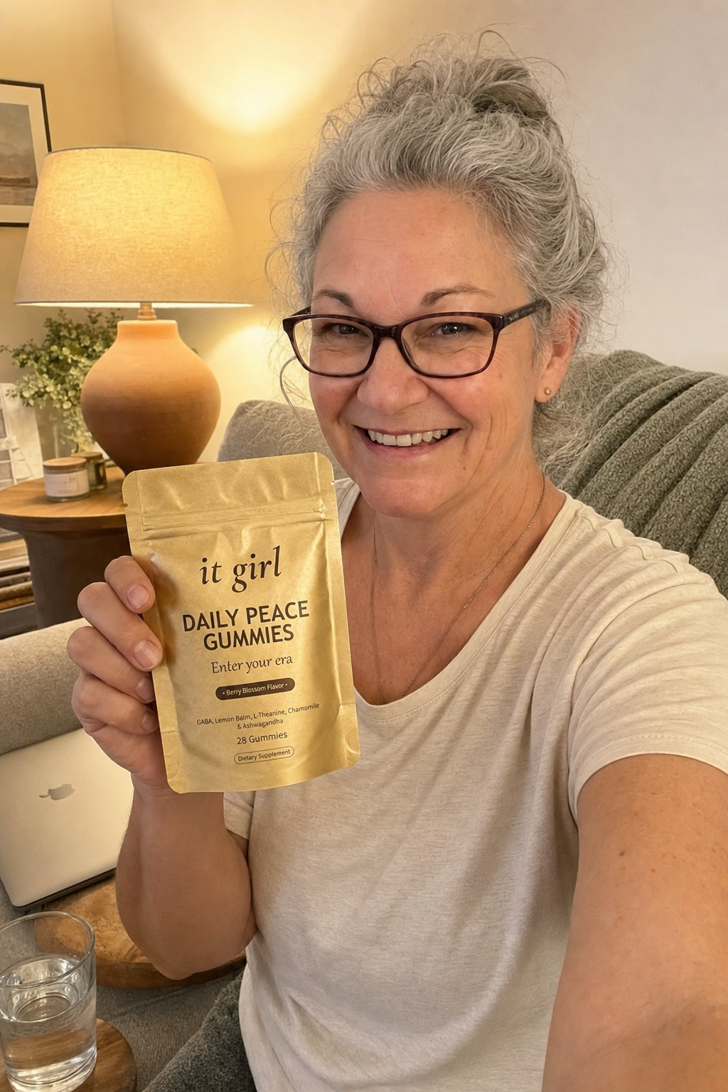 Composed evening selfie of a woman about sixty on her couch at 7:43, laptop closed, holding the gold It Girl Daily Peace pouch, a glass of water nearby, no wine in sight