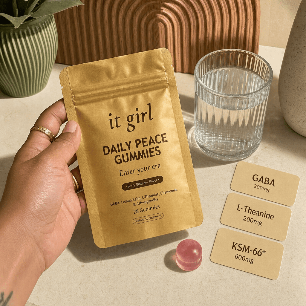 Warm product reveal of the gold It Girl Daily Peace pouch beside a single pink gummy and three dose cards reading GABA 200mg, L-Theanine 200mg, KSM-66 600mg, the full studied stack
