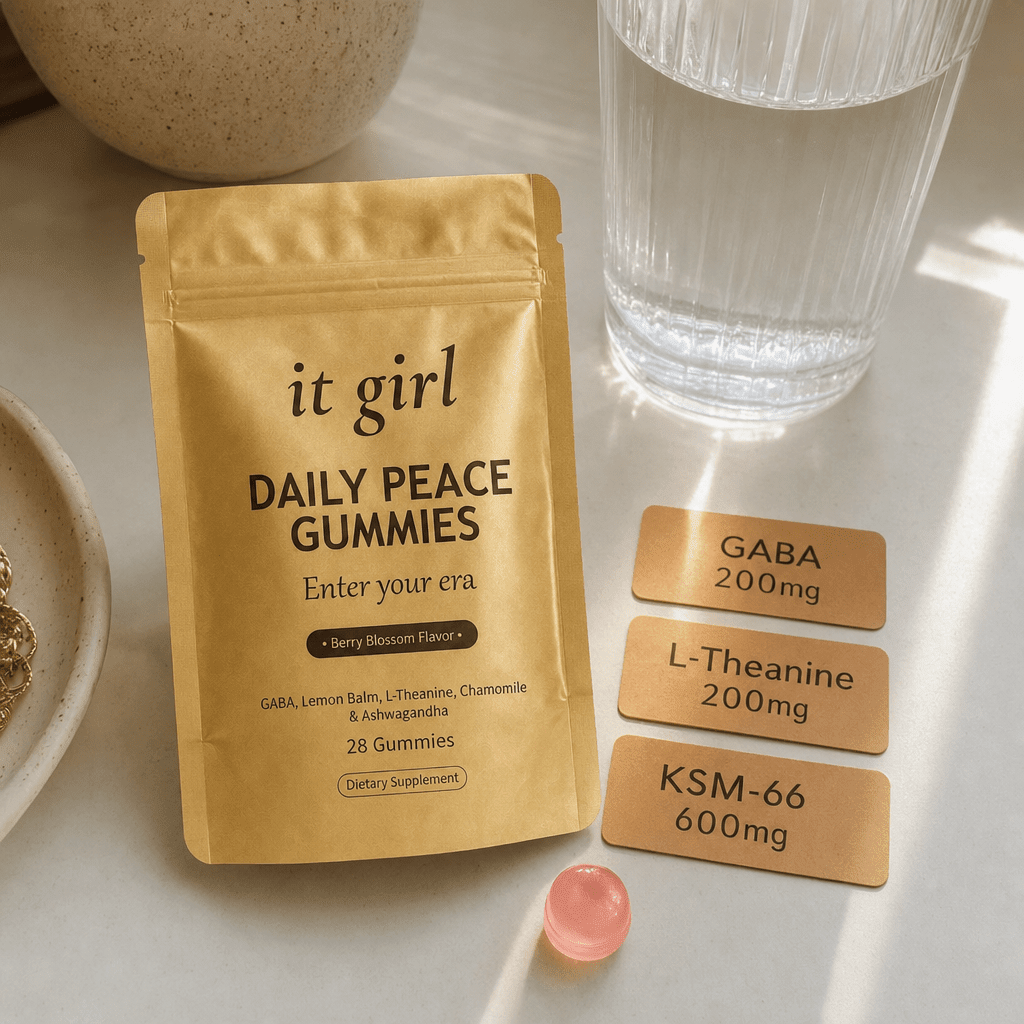 Warm product reveal of the gold It Girl Daily Peace pouch beside a single pink gummy and three dose cards reading GABA 200mg, L-Theanine 200mg, KSM-66 600mg on a cream surface, the full studied stack