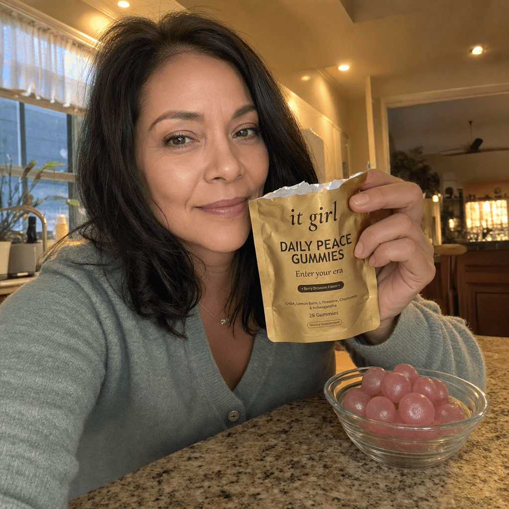 Real photo of a calm put-together woman in her late forties at home holding the soft-gold It Girl Daily Peace pouch with its top torn open toward the camera, a glass bowl of pink gummies beside her, warm relaxed evening light