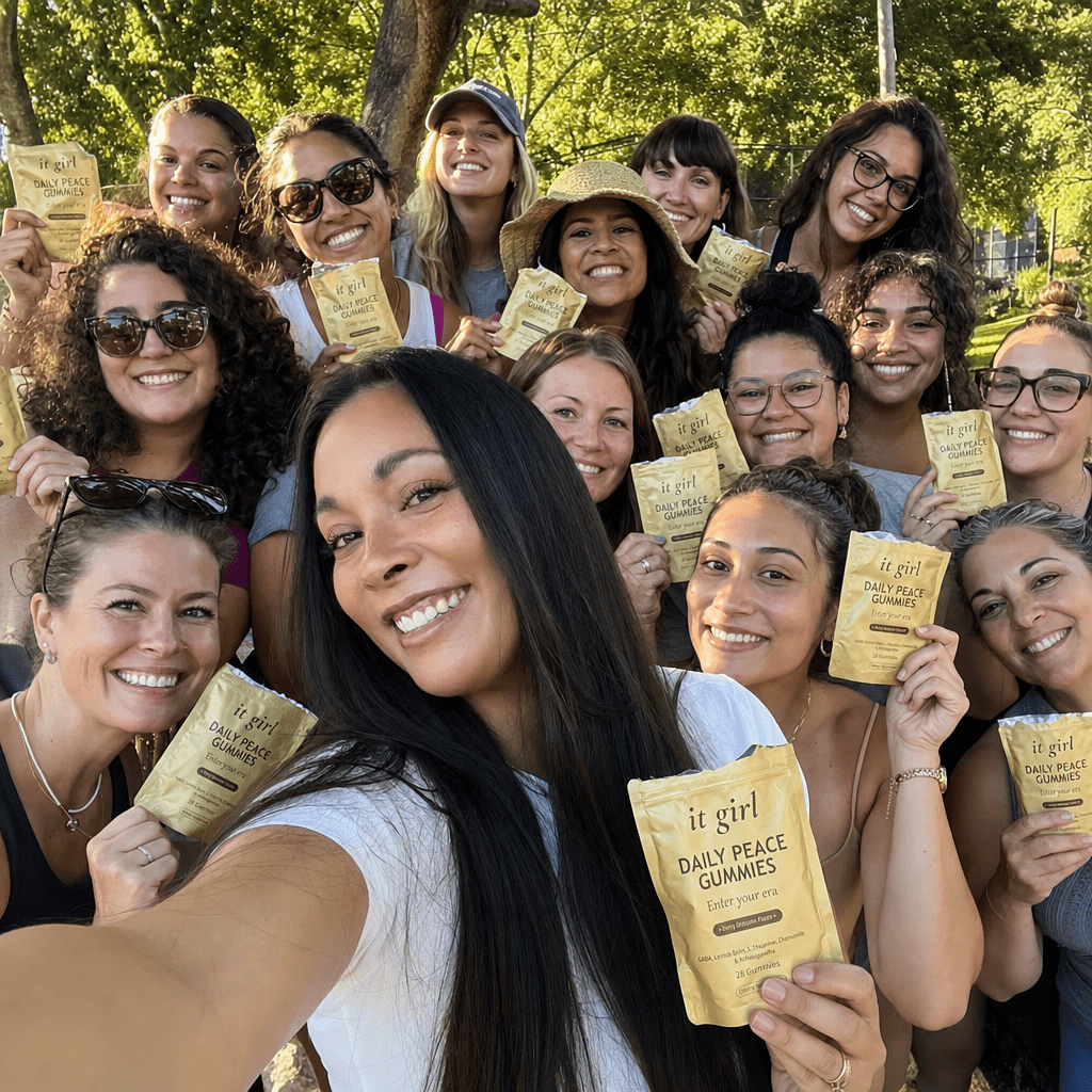 Real group selfie of more than a dozen happy women of different ages crowded together outdoors in warm light, several holding up soft-gold It Girl Daily Peace pouches that are all torn open at the top, candid celebratory community photo