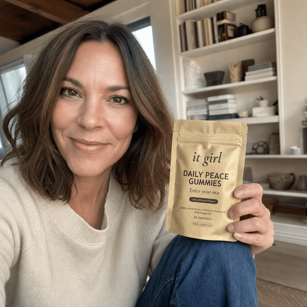 Real photo of a warm professional woman in her late forties with shoulder-length brown hair, a Women's Mental Wellness contributor, sitting in a bright room holding the soft-gold It Girl Daily Peace pouch and smiling at the camera, trusting and calm