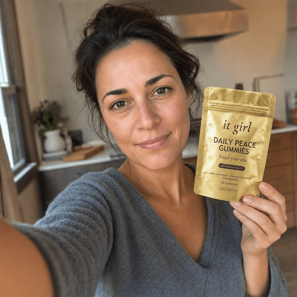 Candid first-person selfie of a working mom about thirty-eight holding her phone at arm's length at a natural off-center angle in a lived-in kitchen, done-up but real not airbrushed, holding the matte-gold It Girl Daily Peace pouch up with the front label facing camera, the product reveal she found